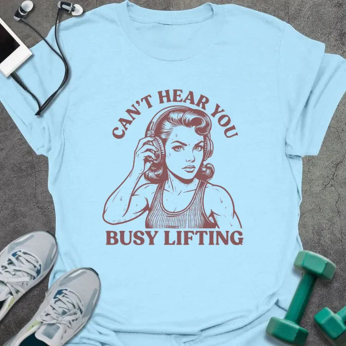 Light blue t-shirt printed with "Can't Hear You Busy Lifting" in rust, with an illustration of a woman with headphones