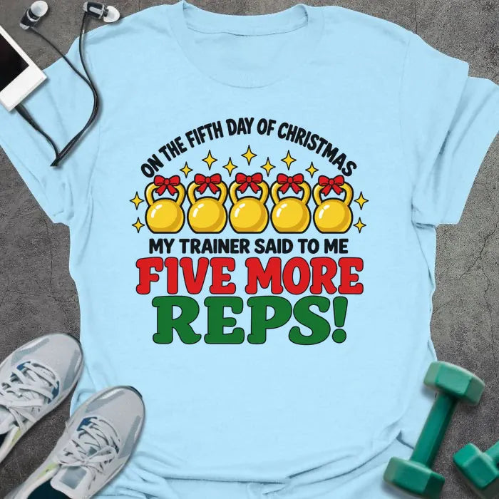 Light blue t-shirt printed with "ON THE FIFTH DAY OF CHRISTMAS MY TRAINER SAID TO ME, FIVE MORE REPS!" in black, red and green, with five gold kettlebells tied with red bows