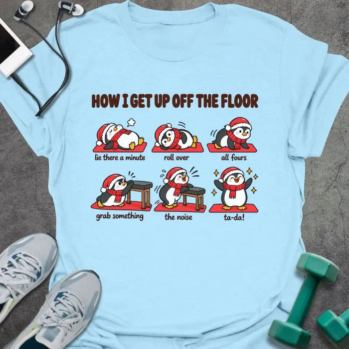 Light blue t-shirt printed with "HOW I GET UP OFF THE FLOOR" in red, with six penguins in Santa hats showing the steps: lie there a minute, roll over, all fours, grab something, the noise, ta-da!