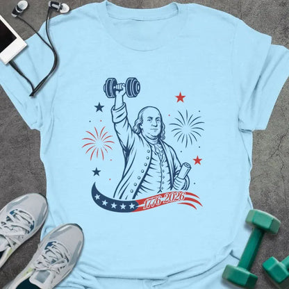 Light blue t-shirt printed with "1776-2026" in navy, red, with Benjamin Franklin holding a dumbbell overhead and a scroll, fireworks and stars