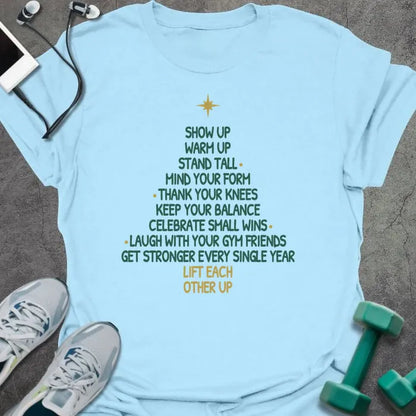 Light blue t-shirt printed with a Christmas tree made of words, "SHOW UP, WARM UP, STAND TALL, MIND YOUR FORM, THANK YOUR KNEES, KEEP YOUR BALANCE, CELEBRATE SMALL WINS, LAUGH WITH YOUR GYM FRIENDS, GET STRONGER EVERY SINGLE YEAR, LIFT EACH OTHER UP", in green and gold with a gold star on top