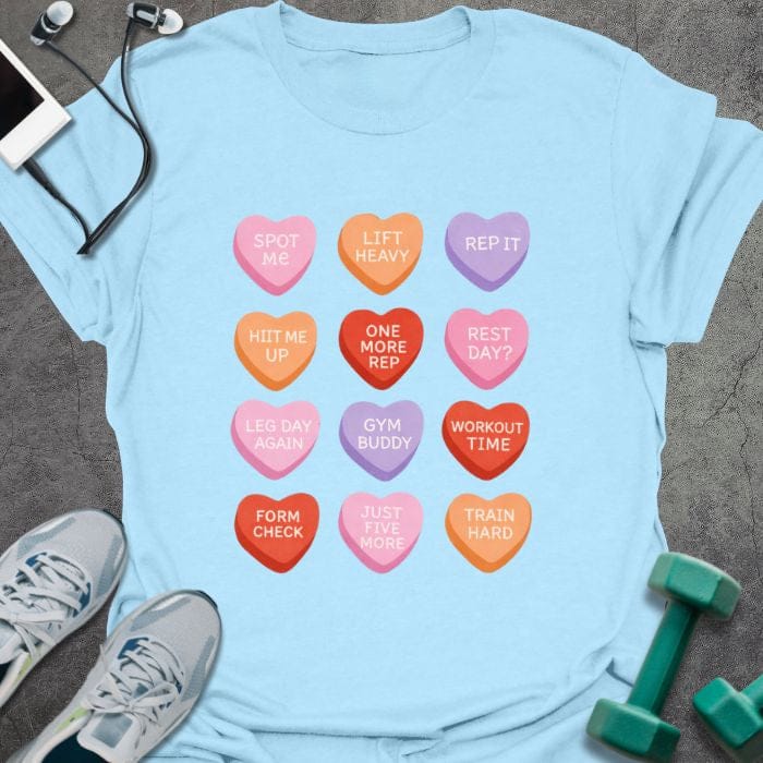 Light blue t-shirt printed with "Spot Me Lift Heavy Rep it HIIT Me Up One More Rep Rest Day? Leg Day Again Gym Buddy Workout Time Form Check Just Five More Train Hard" in pink, red, orange, purple, with a 4x3 grid of colorful candy hearts