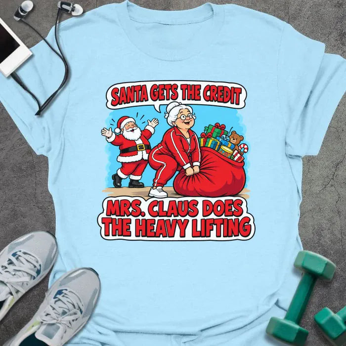 Light blue t-shirt printed with "SANTA GETS THE CREDIT, MRS. CLAUS DOES THE HEAVY LIFTING" in red and white, with Mrs. Claus hauling a sack of gifts while Santa waves