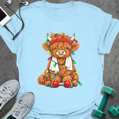 Light blue t-shirt printed with a fluffy highland cow in a red headband and towel, tangled in Christmas lights beside a barbell with ornament plates