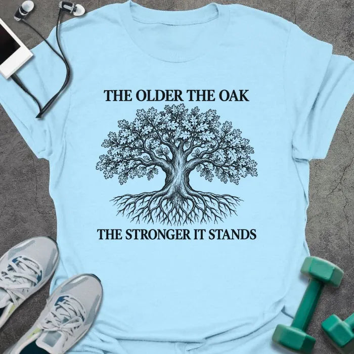 Light blue t-shirt printed with "THE OLDER THE OAK, THE STRONGER IT STANDS" in black, with a drawing of an oak tree and its roots