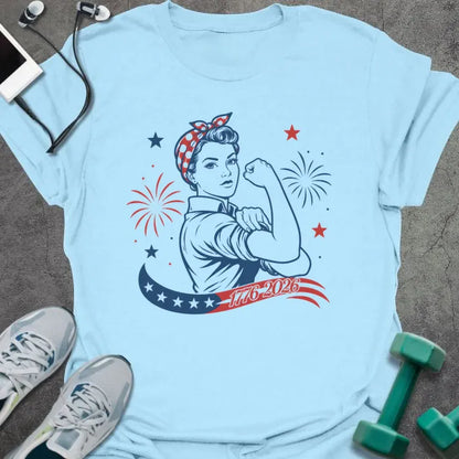 Light blue t-shirt printed with "1776-2026" in navy, red, with Rosie the Riveter flexing her bicep among fireworks and stars