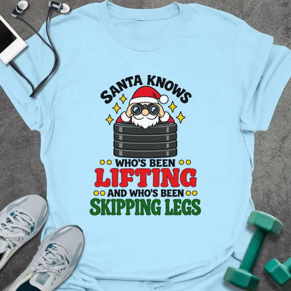 Light blue t-shirt printed with "SANTA KNOWS WHO'S BEEN LIFTING AND WHO'S BEEN SKIPPING LEGS" in black, red and green, with Santa peering through binoculars over a stack of weight plates