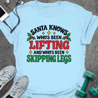Light blue t-shirt printed with "SANTA KNOWS WHO'S BEEN LIFTING AND WHO'S BEEN SKIPPING LEGS" in black, red and green, with holly and gold stars