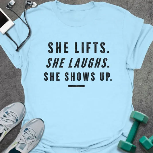 Light Blue t-shirt printed with "SHE LIFTS. SHE LAUGHS. SHE SHOWS UP." in black block lettering with underline