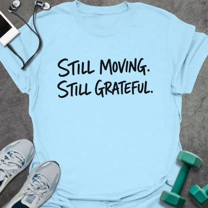 Light Blue t-shirt printed with "Still Moving. Still Grateful." in black handwritten lettering