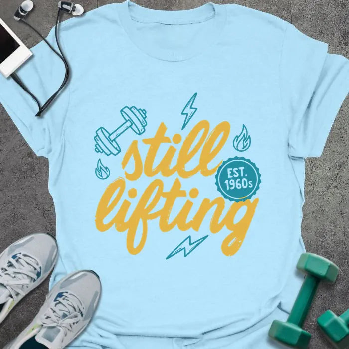 Light blue t-shirt printed with "still lifting EST. 1960s" in yellow and cyan, with lightning bolts and dumbbell graphics