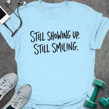 Light Blue t-shirt printed with "STILL SHOWING UP. STILL SMILING." in black handwritten lettering