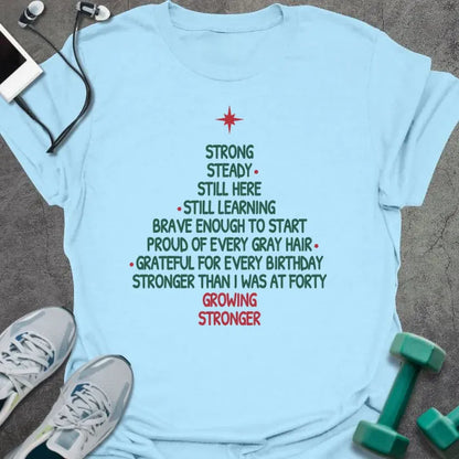 Light blue t-shirt printed with a Christmas tree made of words, "STRONG, STEADY, STILL HERE, STILL LEARNING, BRAVE ENOUGH TO START, PROUD OF EVERY GRAY HAIR, GRATEFUL FOR EVERY BIRTHDAY, STRONGER THAN I WAS AT FORTY, GROWING STRONGER", in green and red with a red star on top