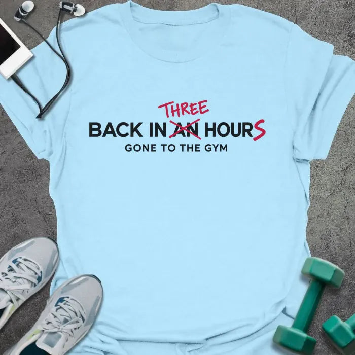 Light blue t-shirt printed with "BACK IN AN HOUR, GONE TO THE GYM" in black, with "AN" crossed out and "THREE" and an extra S added in red to read back in three hours