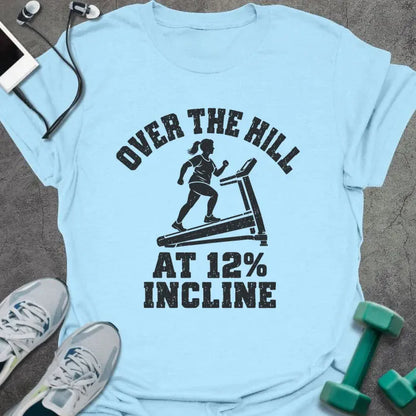 Light blue t-shirt printed with "OVER THE HILL AT 12% INCLINE" in charcoal, with a woman walking on a treadmill