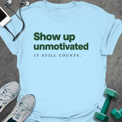 Light blue t-shirt printed with "Show up unmotivated IT STILL COUNTS." in green and black