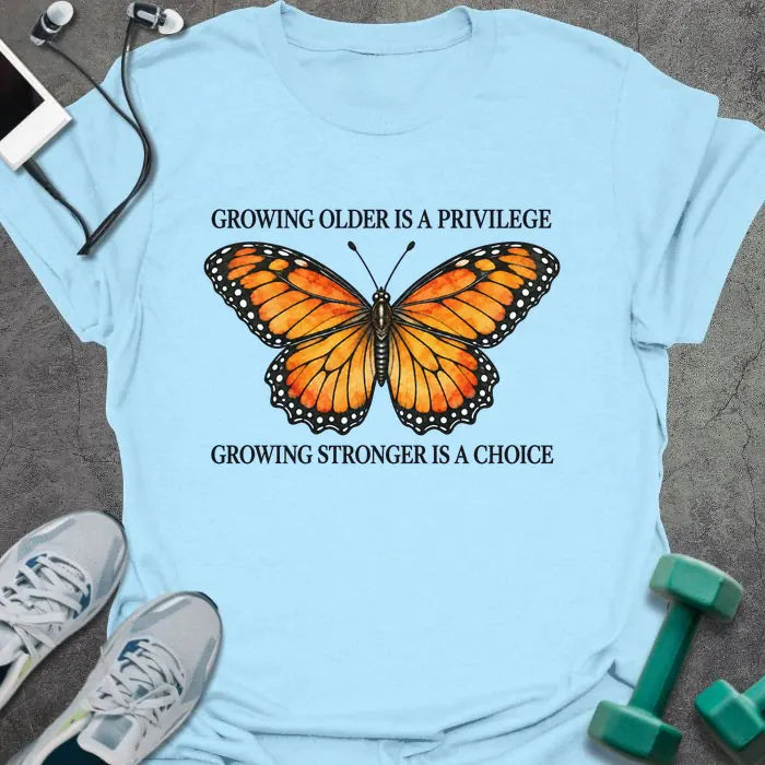 Light blue t-shirt printed with "GROWING OLDER IS A PRIVILEGE, GROWING STRONGER IS A CHOICE" in black, with an orange watercolor monarch butterfly