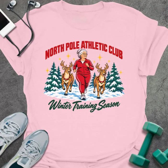 Light pink t-shirt printed with "NORTH POLE ATHLETIC CLUB" in red and "Winter Training Season" in green script, with a white-haired woman in a red tracksuit jogging between two reindeer through snowy pine trees