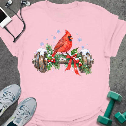 Light pink t-shirt printed with a watercolor red cardinal perched on a snowy dumbbell trimmed with holly, pine branches and a red bow, with blue snowflakes
