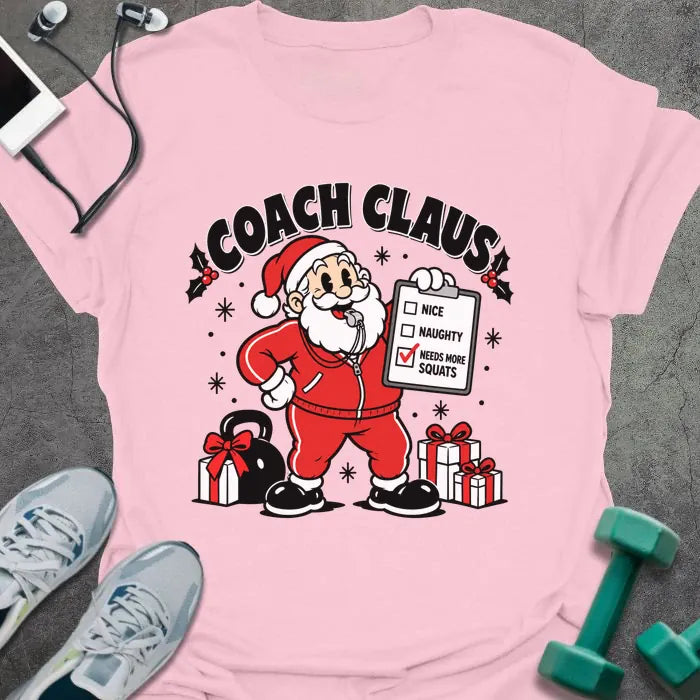 Light pink t-shirt printed with "COACH CLAUS" in black, with a cartoon Santa in a red tracksuit and whistle holding a clipboard with "NEEDS MORE SQUATS" checked, next to a kettlebell and wrapped presents