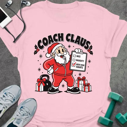 Light pink t-shirt printed with "COACH CLAUS" in black, with a cartoon Santa in a red tracksuit and whistle holding a clipboard with "NEEDS MORE SQUATS" checked, next to a kettlebell and wrapped presents
