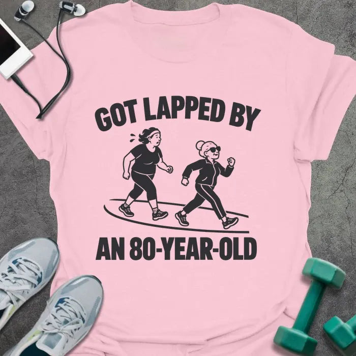 Light pink t-shirt printed with "GOT LAPPED BY AN 80-YEAR-OLD" in black, with a tired jogger being passed by an older woman in sunglasses speed walking on a track