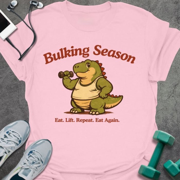 Light pink t-shirt printed with "Bulking Season Eat. Lift. Repeat. Eat Again." in brown and green, with dinosaur lifting weights graphic
