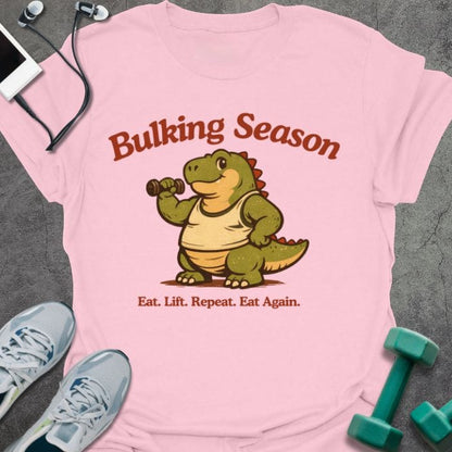 Light pink t-shirt printed with "Bulking Season Eat. Lift. Repeat. Eat Again." in brown and green, with dinosaur lifting weights graphic