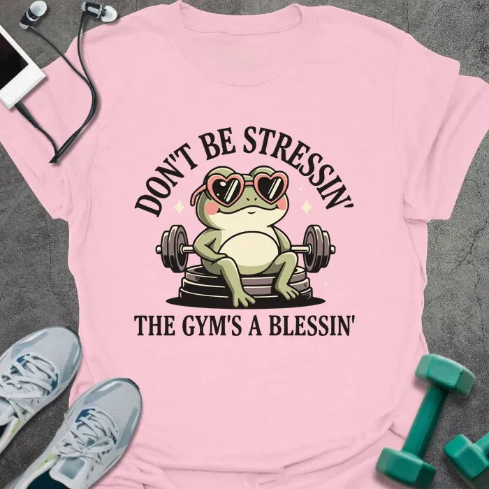 Light pink t-shirt printed with "Don't Be Stressin' the Gym's a Blessin'" in red, with an illustration of a frog with heart-shaped sunglasses lifting dumbbells
