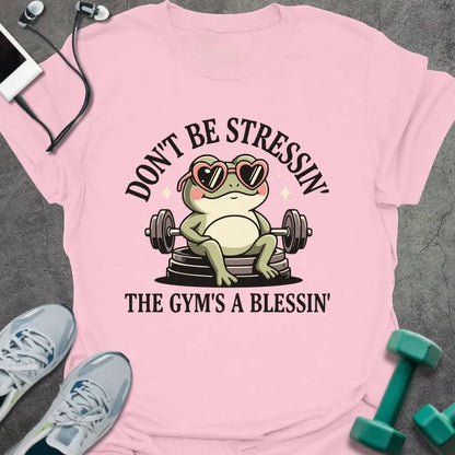 Light pink t-shirt printed with "Don't Be Stressin' the Gym's a Blessin'" in red, with an illustration of a frog with heart-shaped sunglasses lifting dumbbells