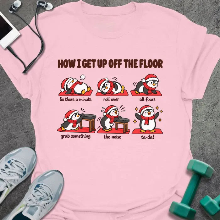 Light pink t-shirt printed with "HOW I GET UP OFF THE FLOOR" in red, with six penguins in Santa hats showing the steps: lie there a minute, roll over, all fours, grab something, the noise, ta-da!
