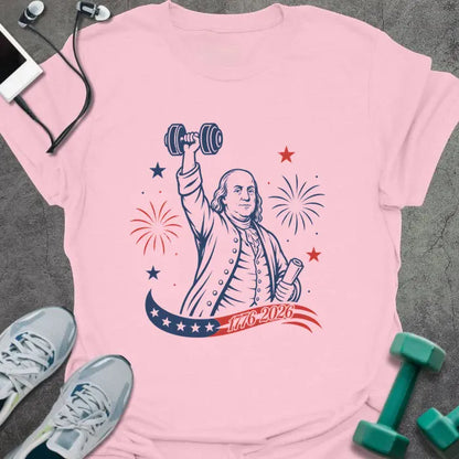 Light pink t-shirt printed with "1776-2026" in navy, red, with Benjamin Franklin holding a dumbbell overhead and a scroll, fireworks and stars