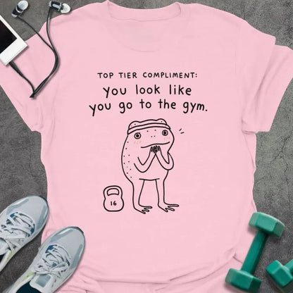 Light Pink t-shirt printed with "TOP TIER COMPLIMENT: you look like you go to the gym." in black, with frog graphic holding barbells