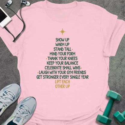 Light pink t-shirt printed with a Christmas tree made of words, "SHOW UP, WARM UP, STAND TALL, MIND YOUR FORM, THANK YOUR KNEES, KEEP YOUR BALANCE, CELEBRATE SMALL WINS, LAUGH WITH YOUR GYM FRIENDS, GET STRONGER EVERY SINGLE YEAR, LIFT EACH OTHER UP", in green and gold with a gold star on top