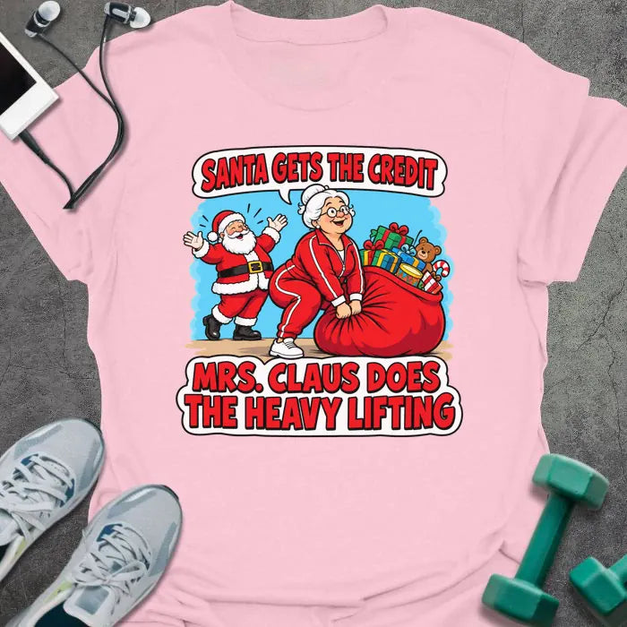 Light pink t-shirt printed with "SANTA GETS THE CREDIT, MRS. CLAUS DOES THE HEAVY LIFTING" in red and white, with Mrs. Claus hauling a sack of gifts while Santa waves