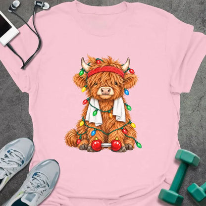 Light pink t-shirt printed with a fluffy highland cow in a red headband and towel, tangled in Christmas lights beside a barbell with ornament plates