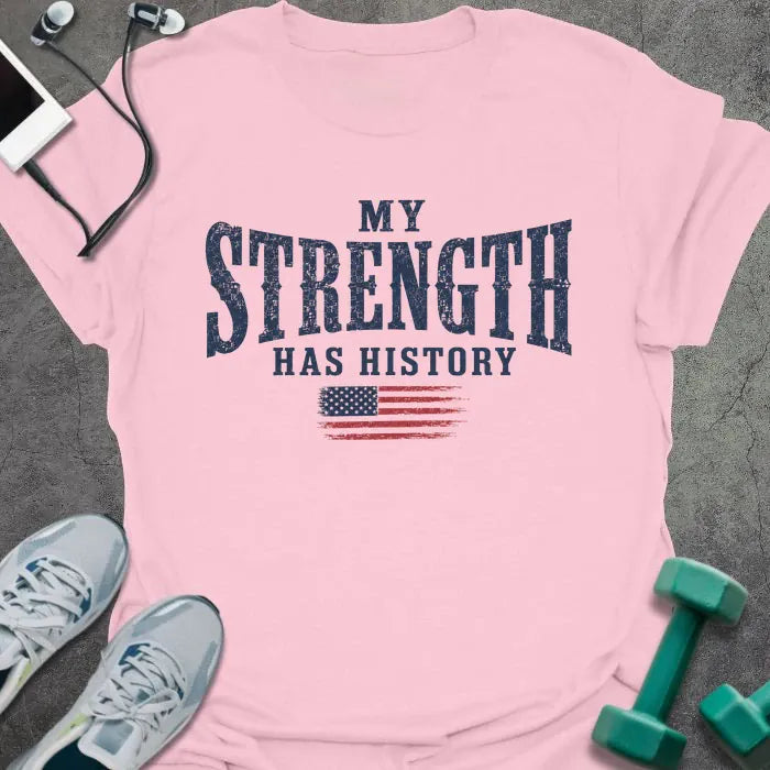 Light pink t-shirt printed with "My Strength Has History" in blue and red, with a distressed American flag
