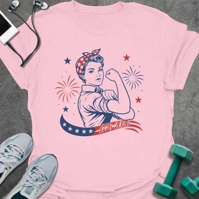 Light pink t-shirt printed with "1776-2026" in navy, red, with Rosie the Riveter flexing her bicep among fireworks and stars