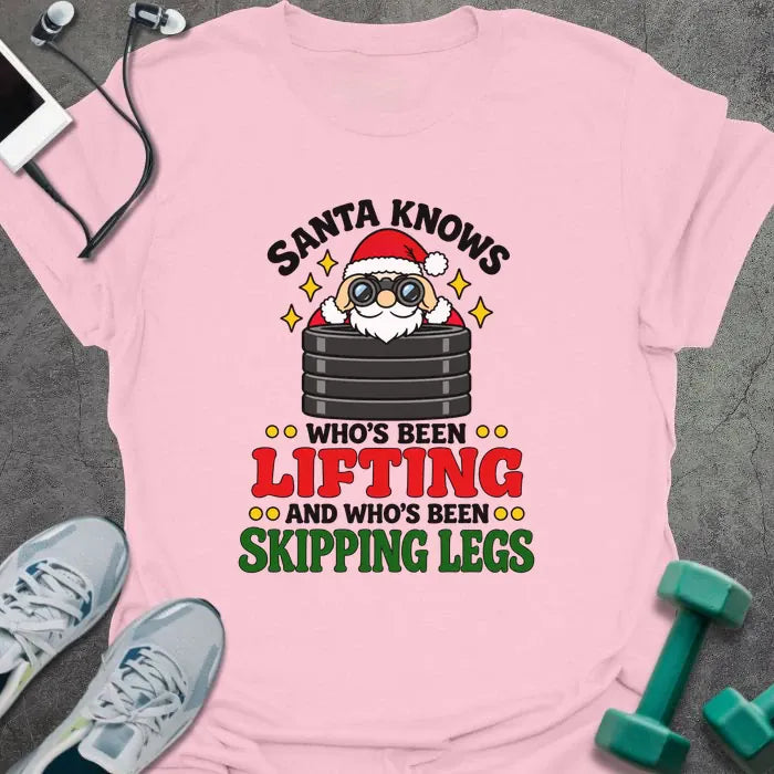 Light pink t-shirt printed with "SANTA KNOWS WHO'S BEEN LIFTING AND WHO'S BEEN SKIPPING LEGS" in black, red and green, with Santa peering through binoculars over a stack of weight plates