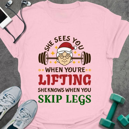 Light pink t-shirt printed with "She Sees You When You're Lifting Sheknows When You Skip Legs" in black, red, and gold, with female Santa and barbell