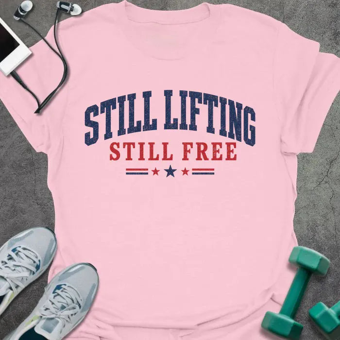 Light pink t-shirt printed with "Still Lifting Still Free" in blue and red, with stars