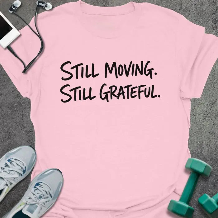 Light Pink t-shirt printed with "Still Moving. Still Grateful." in black handwritten lettering