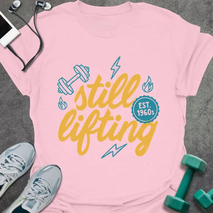 Light pink t-shirt printed with "still lifting EST. 1960s" in yellow and cyan, with lightning bolts and dumbbell graphics