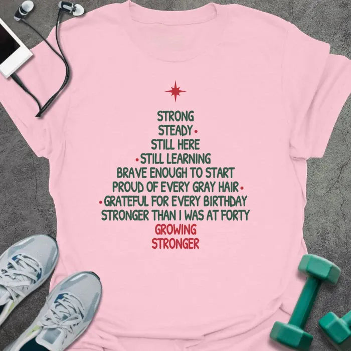 Light pink t-shirt printed with a Christmas tree made of words, "STRONG, STEADY, STILL HERE, STILL LEARNING, BRAVE ENOUGH TO START, PROUD OF EVERY GRAY HAIR, GRATEFUL FOR EVERY BIRTHDAY, STRONGER THAN I WAS AT FORTY, GROWING STRONGER", in green and red with a red star on top
