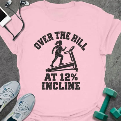 Light pink t-shirt printed with "OVER THE HILL AT 12% INCLINE" in charcoal, with a woman walking on a treadmill
