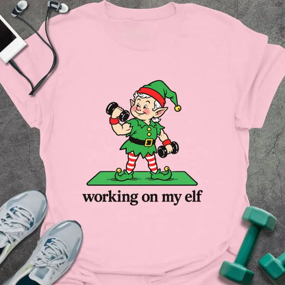 Light pink t-shirt printed with "working on my elf" in black, with a vintage elf lifting dumbbells on a green mat