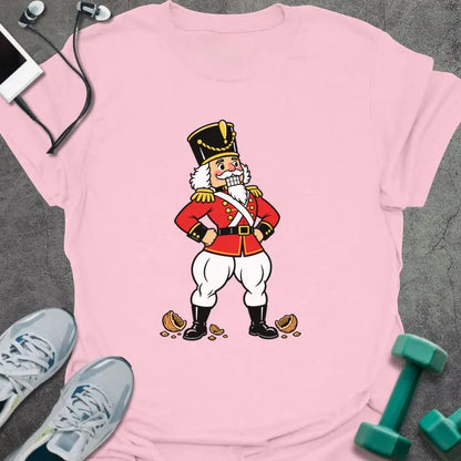 Light pink t-shirt printed with a vintage nutcracker soldier with strong legs, hands on hips, standing over cracked nutshells