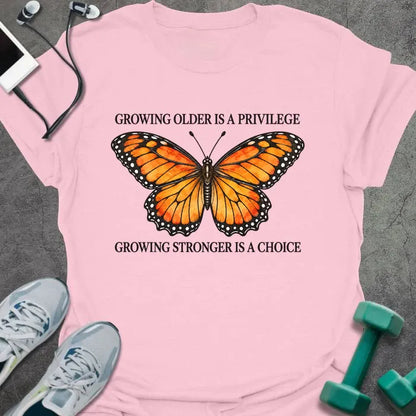 Light pink t-shirt printed with "GROWING OLDER IS A PRIVILEGE, GROWING STRONGER IS A CHOICE" in black, with an orange watercolor monarch butterfly