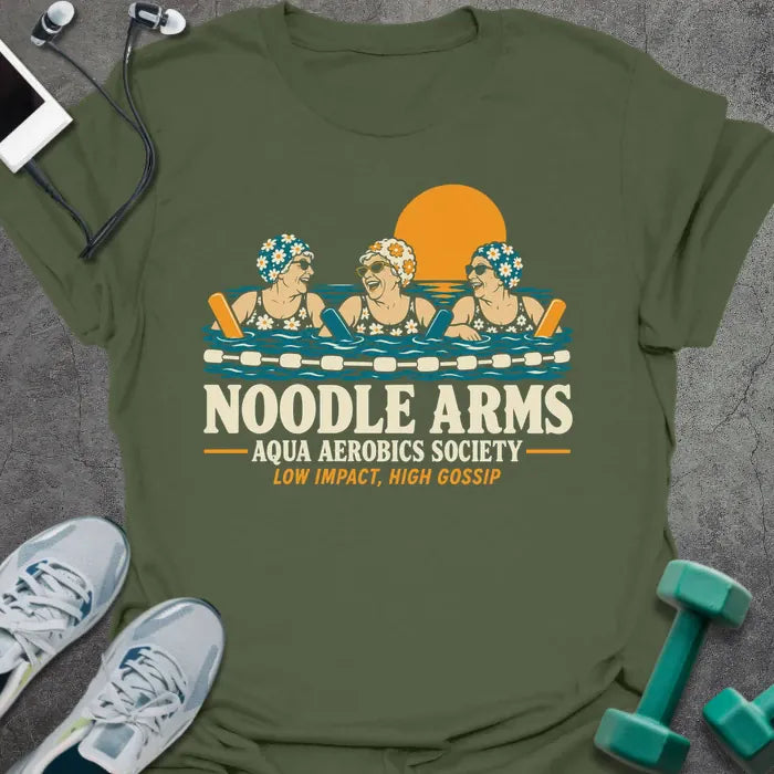 Military green t-shirt printed with "NOODLE ARMS, AQUA AEROBICS SOCIETY, LOW IMPACT, HIGH GOSSIP" in cream and orange, with three older women in flowered swim caps and sunglasses laughing in a pool with pool noodles at sunset