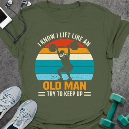 Military green t-shirt printed with "I Know I Lift Like an Old Man Try to Keep Up" in orange, white, green, light blue, with a person lifting and rainbow sunset background
